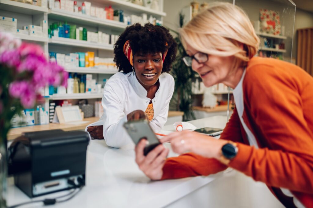Eine Kundin zeigt einer Apothekerin am Tresen den Bildschirm ihres Smartphones.
