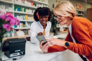 Eine Kundin zeigt einer Apothekerin am Tresen den Bildschirm ihres Smartphones.