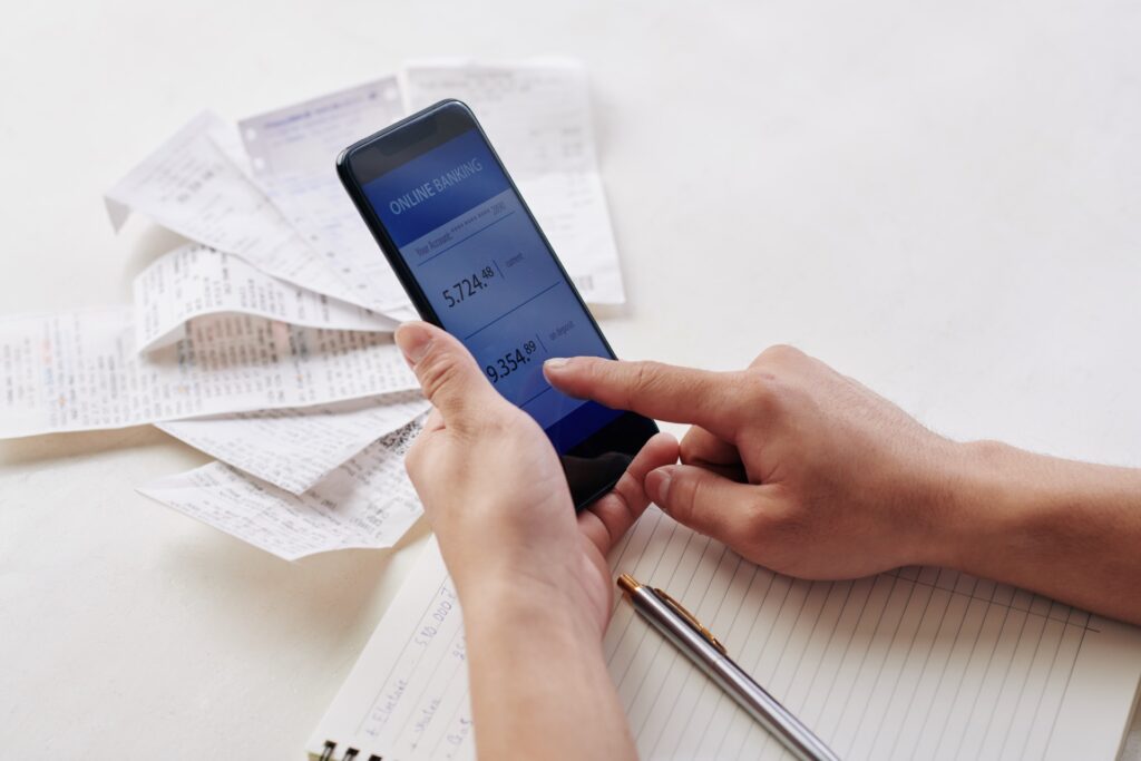 Hand hält ein Smartphone mit geöffneter Banking-App über einem Stapel Rechnungen und Belegen