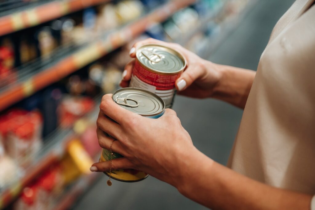 Hände einer Frau halten im Supermarktgang zwei Konservendosen und prüfen sie