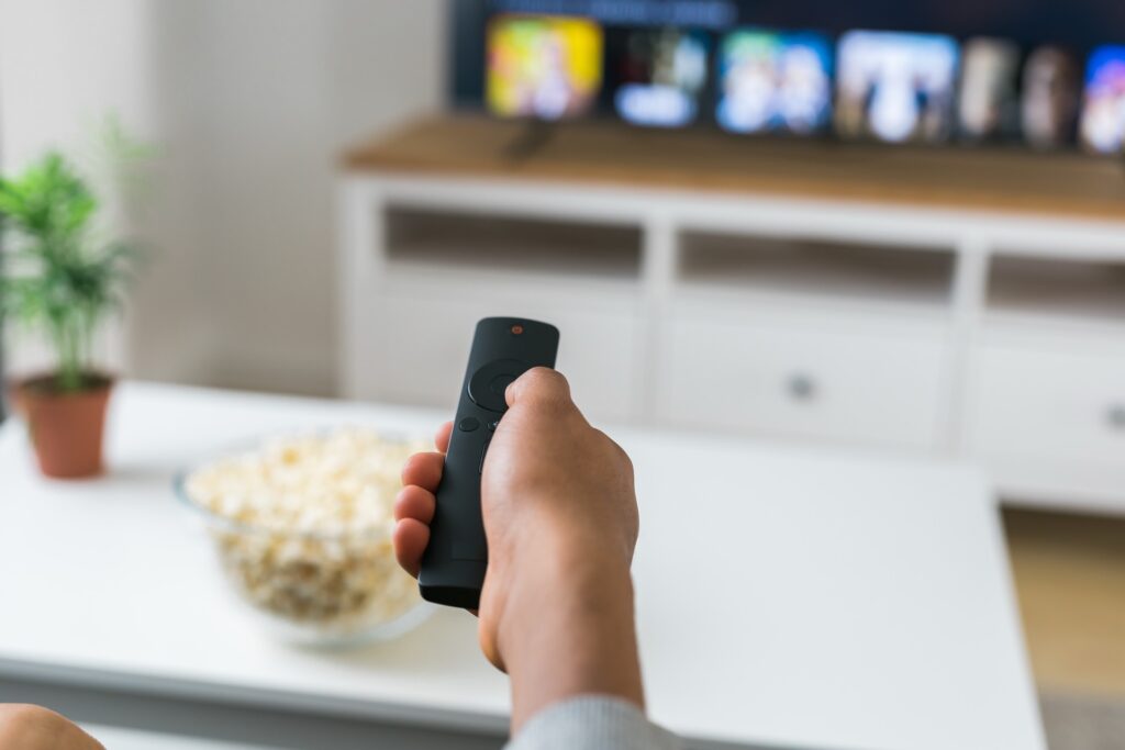 Eine Hand hält eine Fernbedienung vor einen Fernseher mit Streaming-Kacheln, daneben eine Schale Popcorn