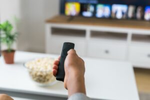 Eine Hand hält eine Fernbedienung vor einen Fernseher mit Streaming-Kacheln, daneben eine Schale Popcorn