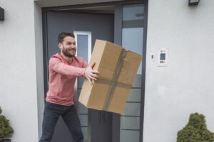 Ein lächelnder Mann trägt einen großen Paketkarton vor einer Haustür