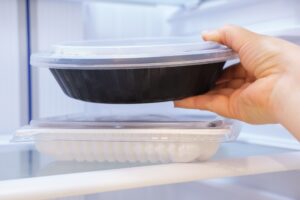 Eine Hand haelt eine runde Plastikdose mit Essen vor einem geoeffneten Kuehlschrank