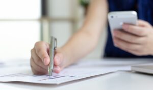 Eine Hand hält ein Smartphone, die andere führt einen Kugelschreiber über ein Dokument auf dem Tisch.