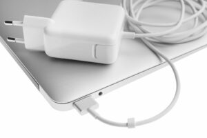 Weißes Netzteil mit Ladekabel liegt auf einem silbernen Laptop, das Kabel steckt seitlich in der Ladebuchse.