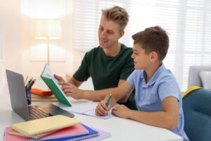 Ein junger Mann hilft einem Jungen am Tisch bei den Hausaufgaben, vor ihnen Laptop, Hefte und ein Buch