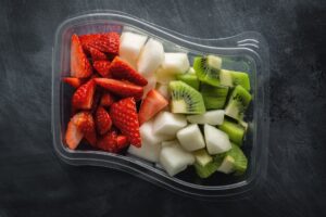 Durchsichtige Plastikschale mit vorgeschnittenen Erdbeeren, Melonen- und Kiwistücken auf dunklem Untergrund, von oben fotografiert