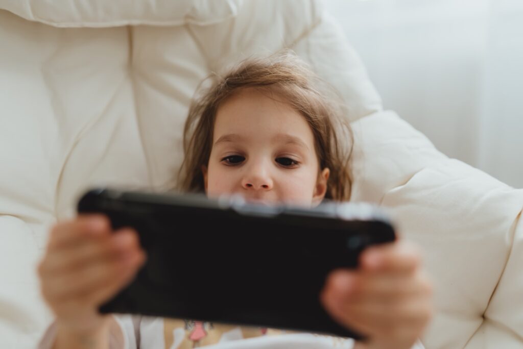 Kleines Mädchen liegt auf einem Kissen und spielt mit einem quer gehaltenen Smartphone