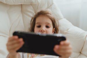 Kleines Mädchen liegt auf einem Kissen und spielt mit einem quer gehaltenen Smartphone