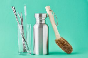 Edelstahl-Trinkflasche neben einer Holzbürste und Metall-Trinkhalmen in einem Glas vor türkisgrünem Hintergrund