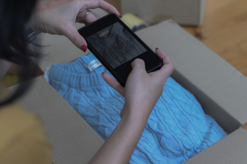 Frau fotografiert mit dem Smartphone einen blauen Strickpullover in einem geöffneten Versandkarton